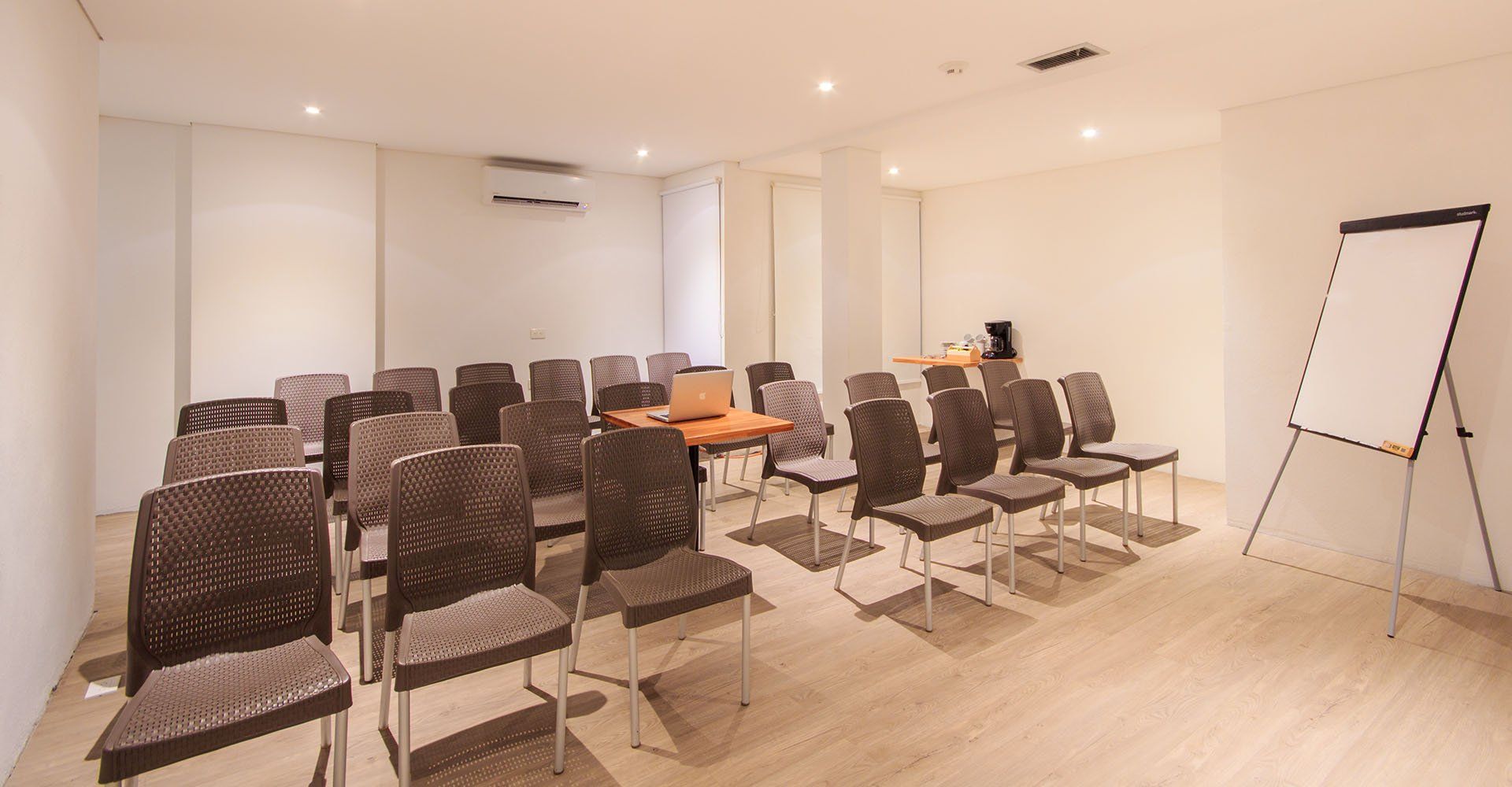 Sala de conferencias vacía con filas de sillas frente a una mesa y una pizarra; colores neutros e iluminación brillante.