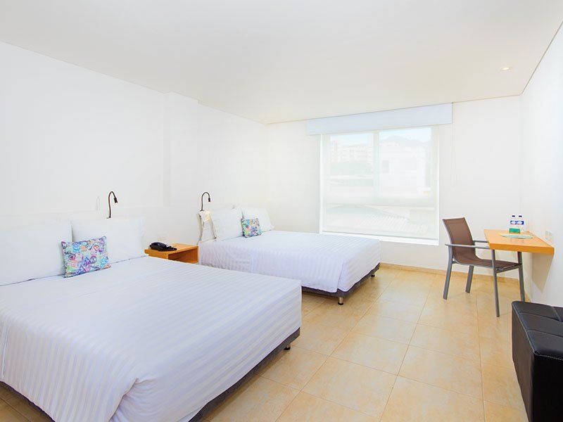 Una habitación de hotel con dos camas blancas, un escritorio y una ventana con persianas blancas. La habitación es mayoritariamente blanca, con suelo color canela.