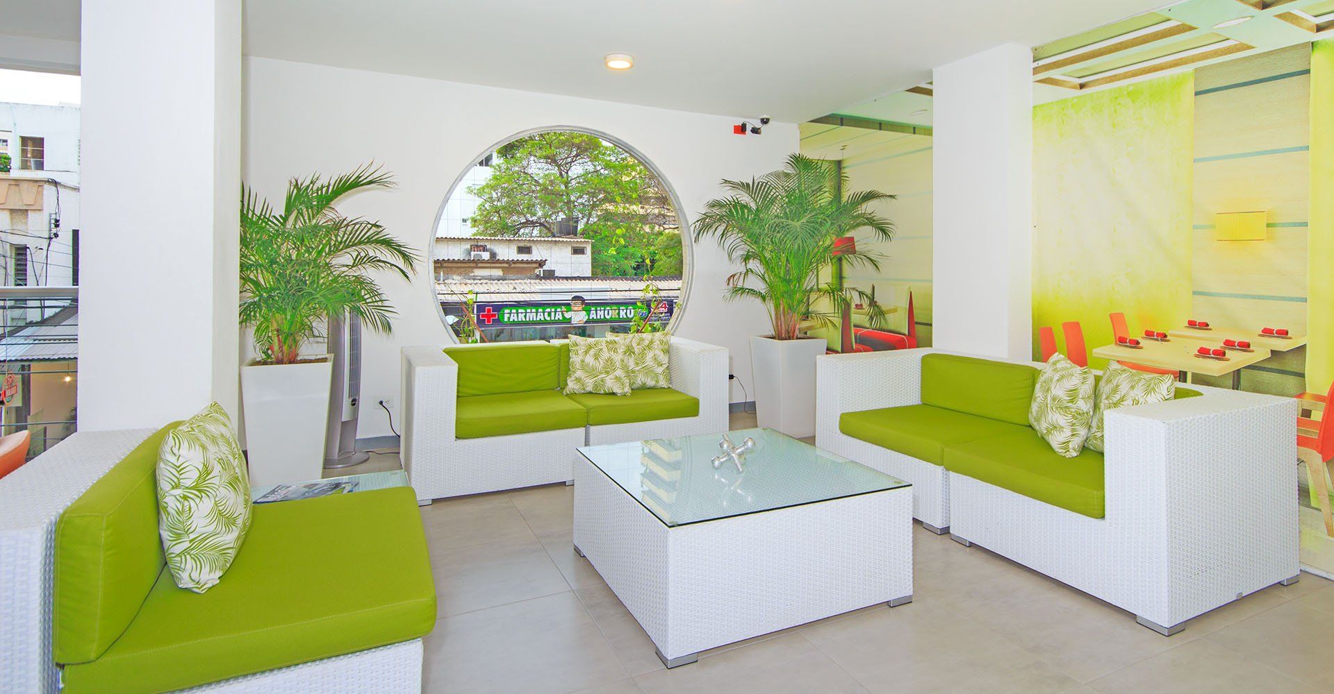 Asientos de mimbre blanco con cojines y almohadas verdes en una sala de estar llena de luz, con una ventana redonda que revela una vista exterior y plantas en macetas.