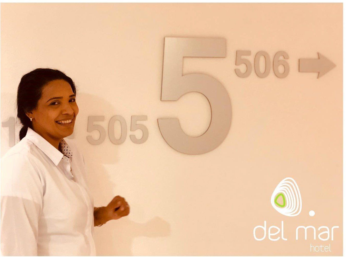 Una mujer sonríe junto a una pared que muestra los números de habitación 505, 5 y 506 con una flecha. El logotipo del 