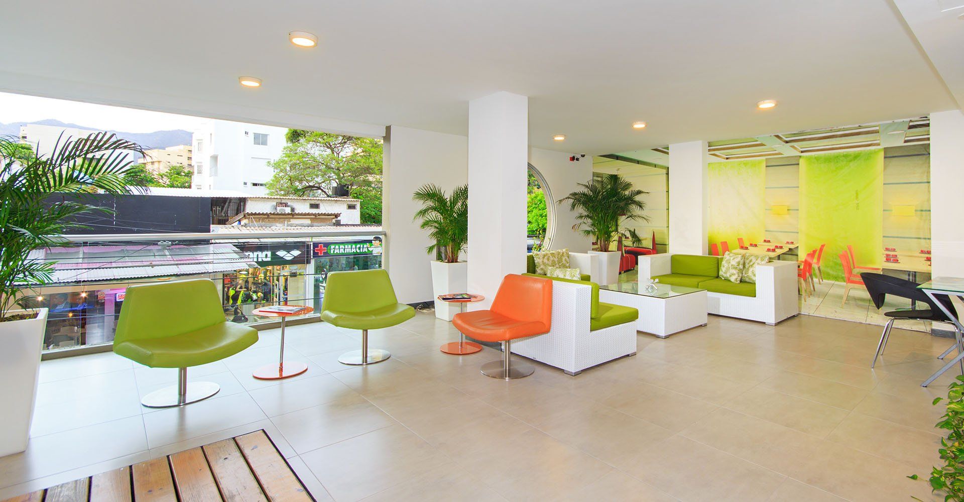 Sala de espera moderna y bien iluminada con asientos verdes y naranjas, paredes blancas y macetas con plantas. La barandilla de cristal da a la calle.