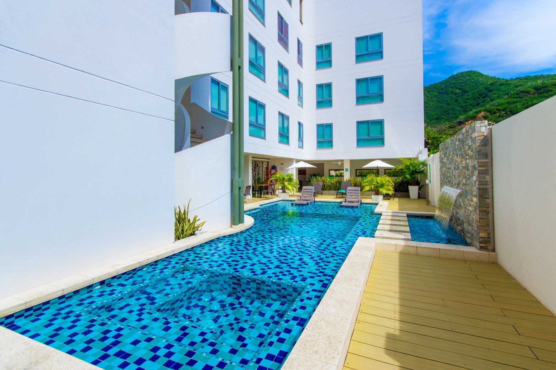 Una piscina de hotel con agua revestida de azulejos azules, rodeada de paredes blancas y un edificio con ventanas verde azulado, con montañas visibles al fondo.