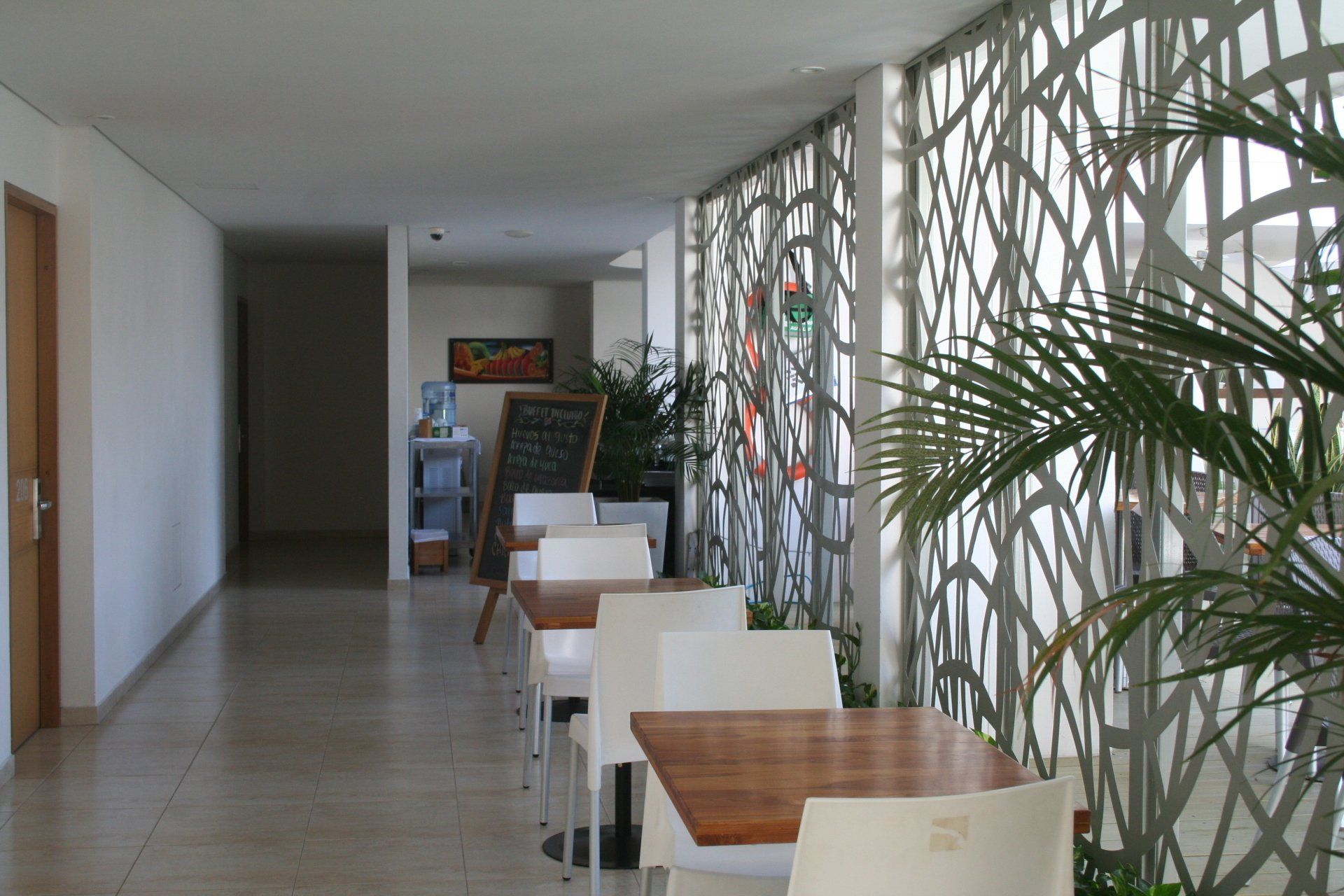 Un pasillo estrecho con mesas y sillas blancas, un biombo decorativo y una planta. El pasillo conduce a una puerta lejana y un letrero de pizarra.