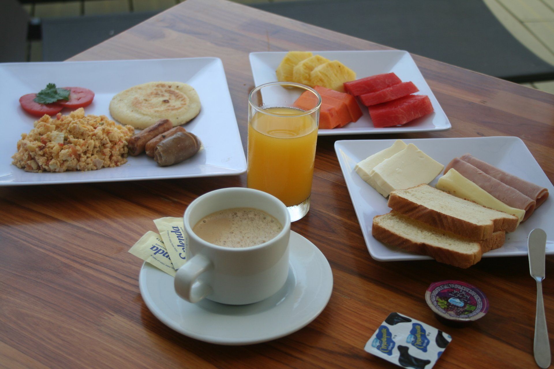 Desayuno servido con café, jugo, fruta, huevos revueltos, arepa, salchicha, queso, jamón y pan en una mesa de madera.