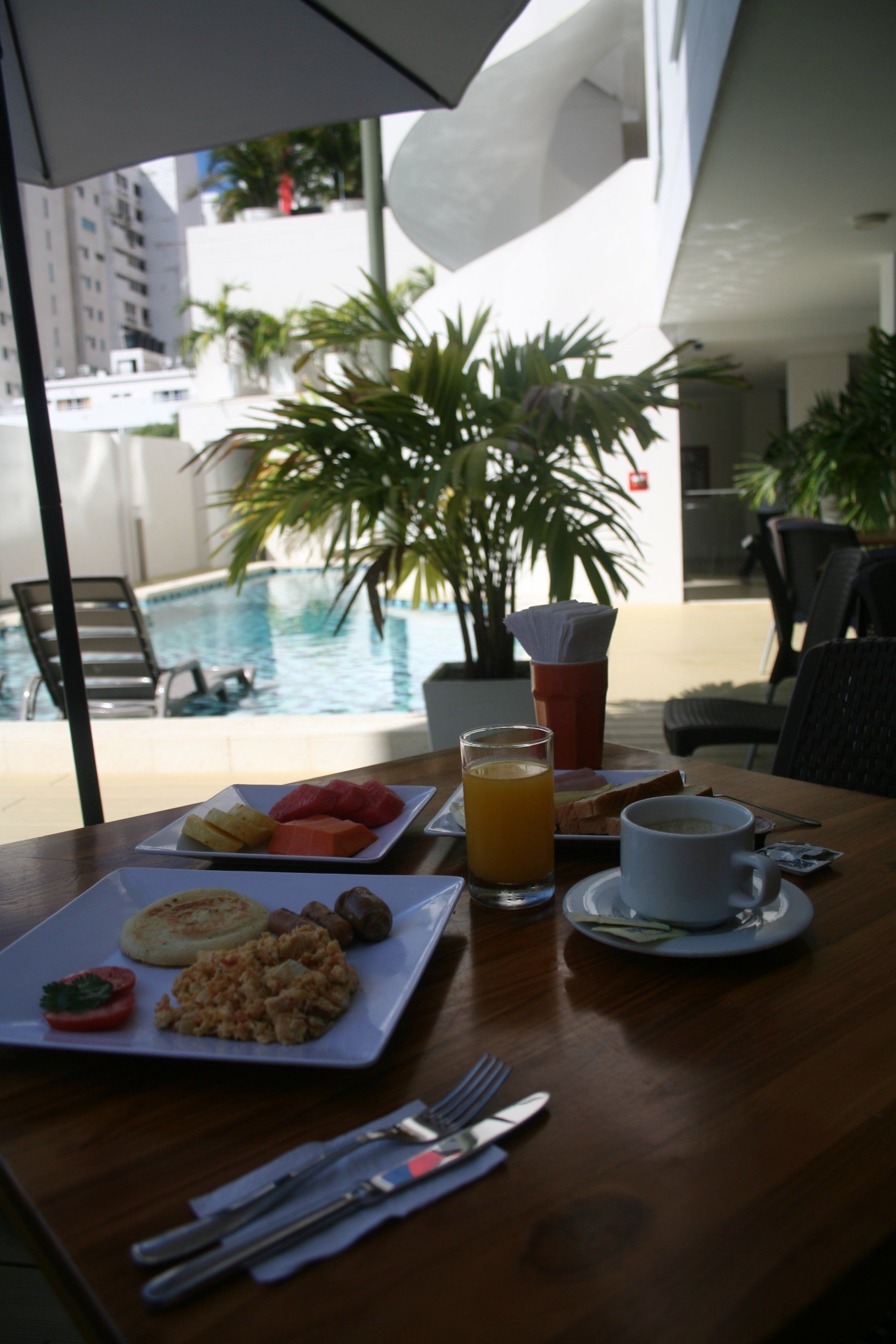 Mesa de desayuno al aire libre junto a la piscina. Comida y bebida en una mesa de madera; incluye un plato de fruta, café y jugo.