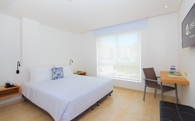 Habitación de hotel con cama, escritorio y ventana. Tiene paredes blancas y suelo de madera clara.