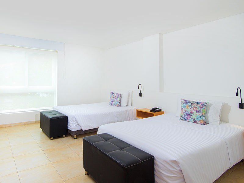 Dos camas individuales en una habitación de hotel blanca, con bancos negros para los pies y almohadas de colores, junto a una ventana.