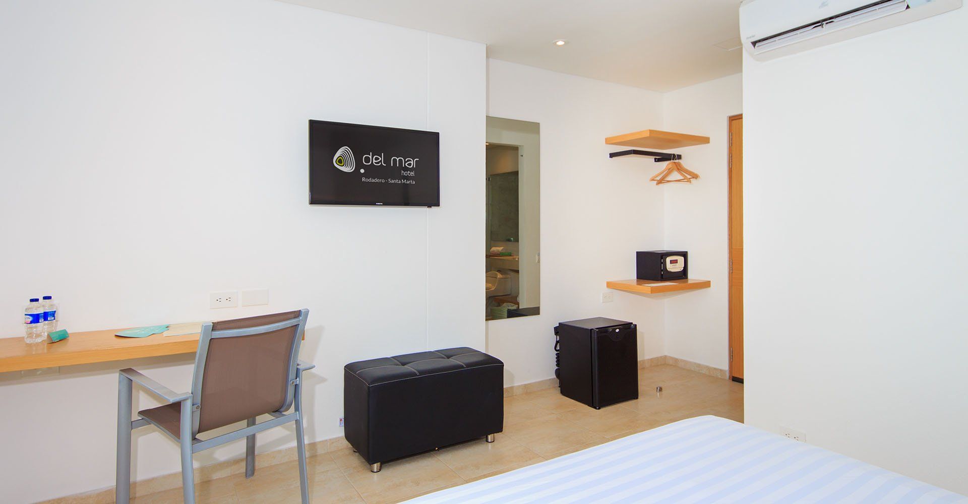 Una habitación de hotel con escritorio, televisión, caja fuerte y un pequeño banco negro con espacio de almacenamiento. Paredes blancas y detalles en madera clara.