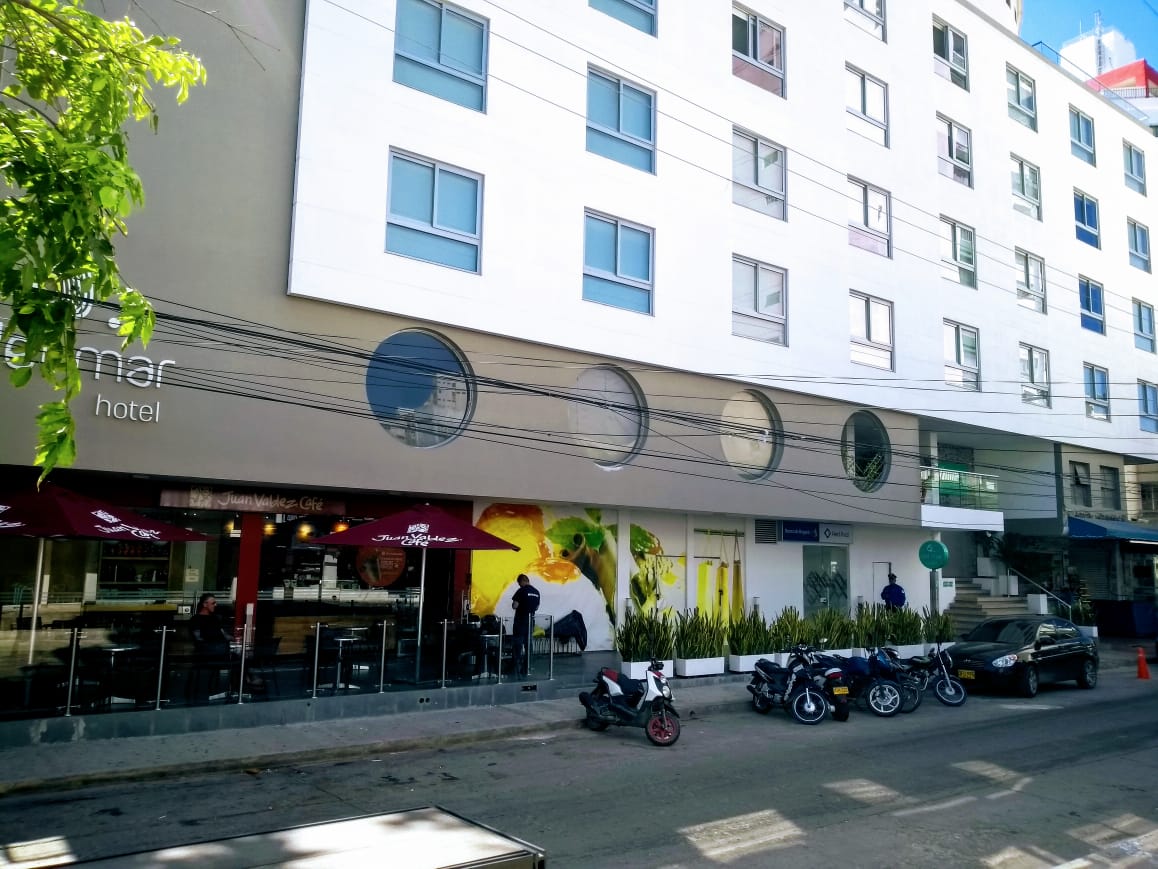 Un hotel blanco con cafetería y motos aparcadas en la calle. La cafetería tiene terraza y un toldo rojo.