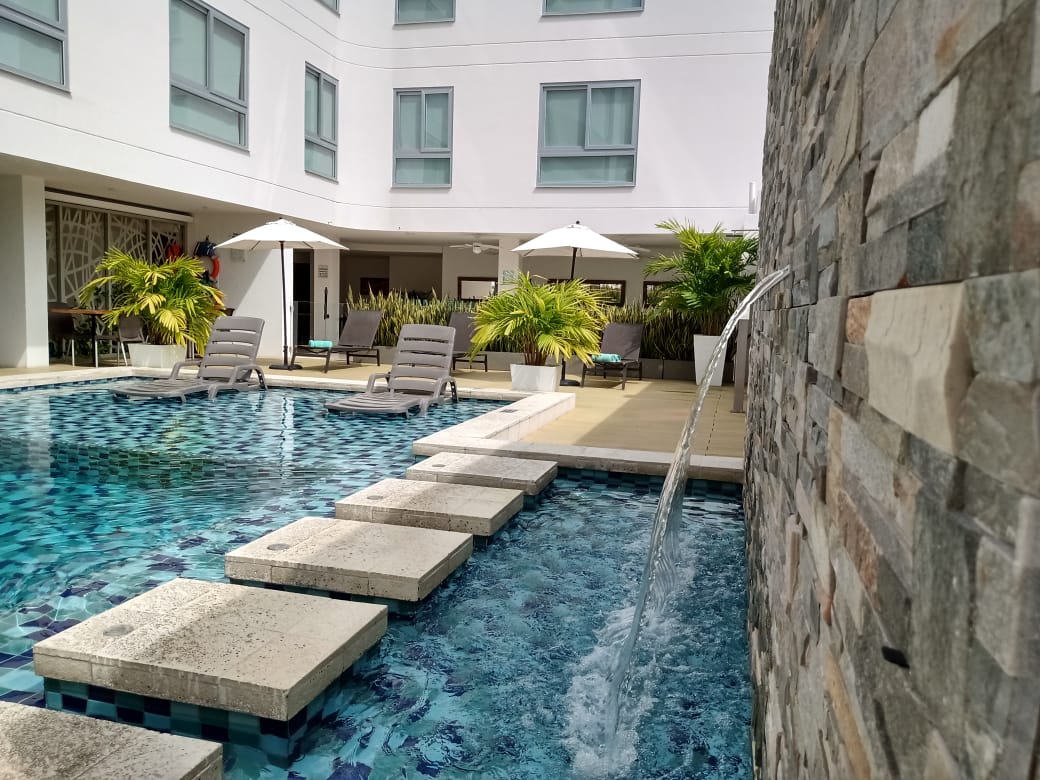 Piscina con escalones, tumbonas y sombrillas frente a un edificio blanco con una fuente de piedra.
