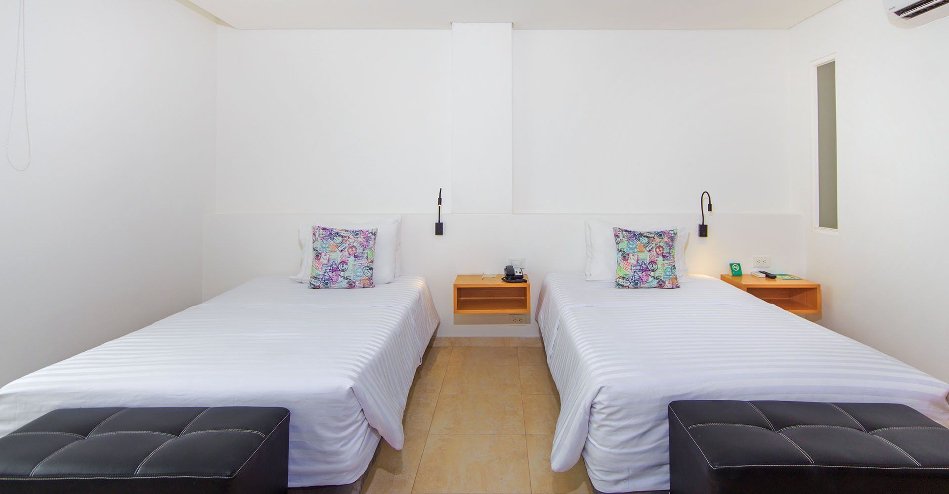 Dos camas individuales en una habitación blanca con mesas de noche a juego, otomanas negras al pie de cada cama y almohadas florales.