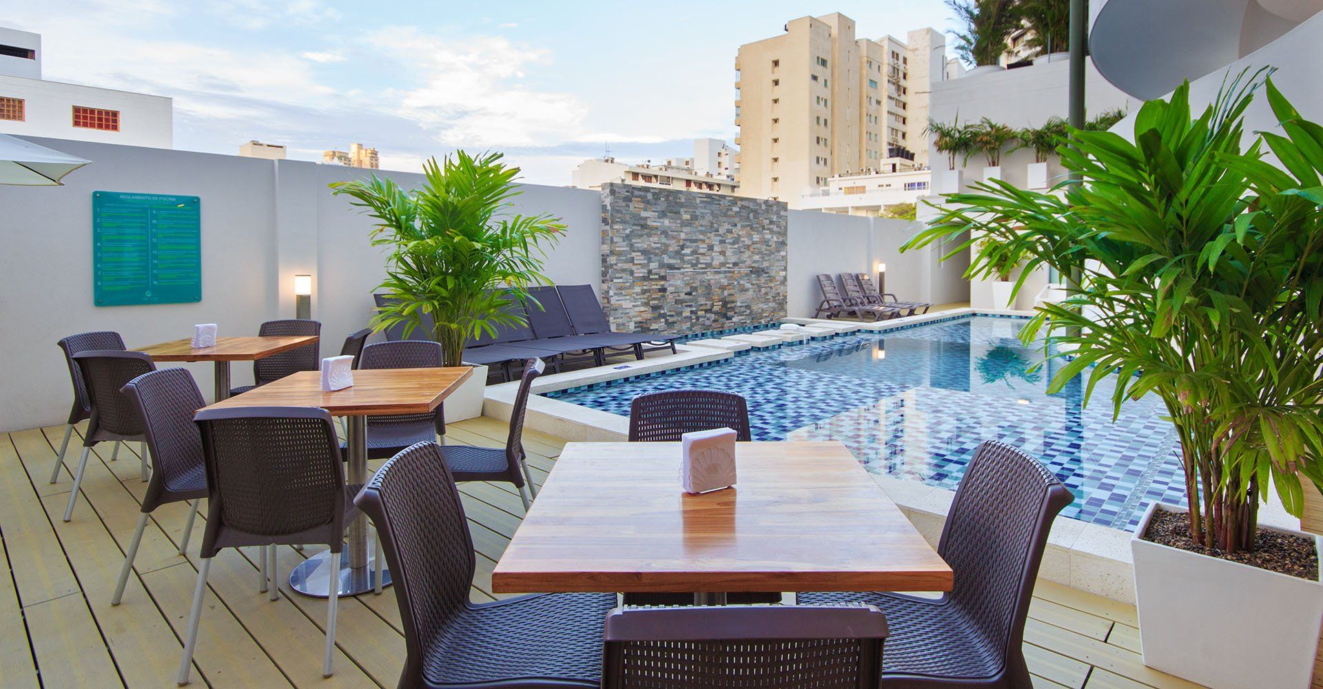 Restaurante al aire libre con vista a la piscina: mesas de madera, sillas oscuras, plantas exuberantes y una piscina de mosaicos azules en un entorno de paredes blancas.