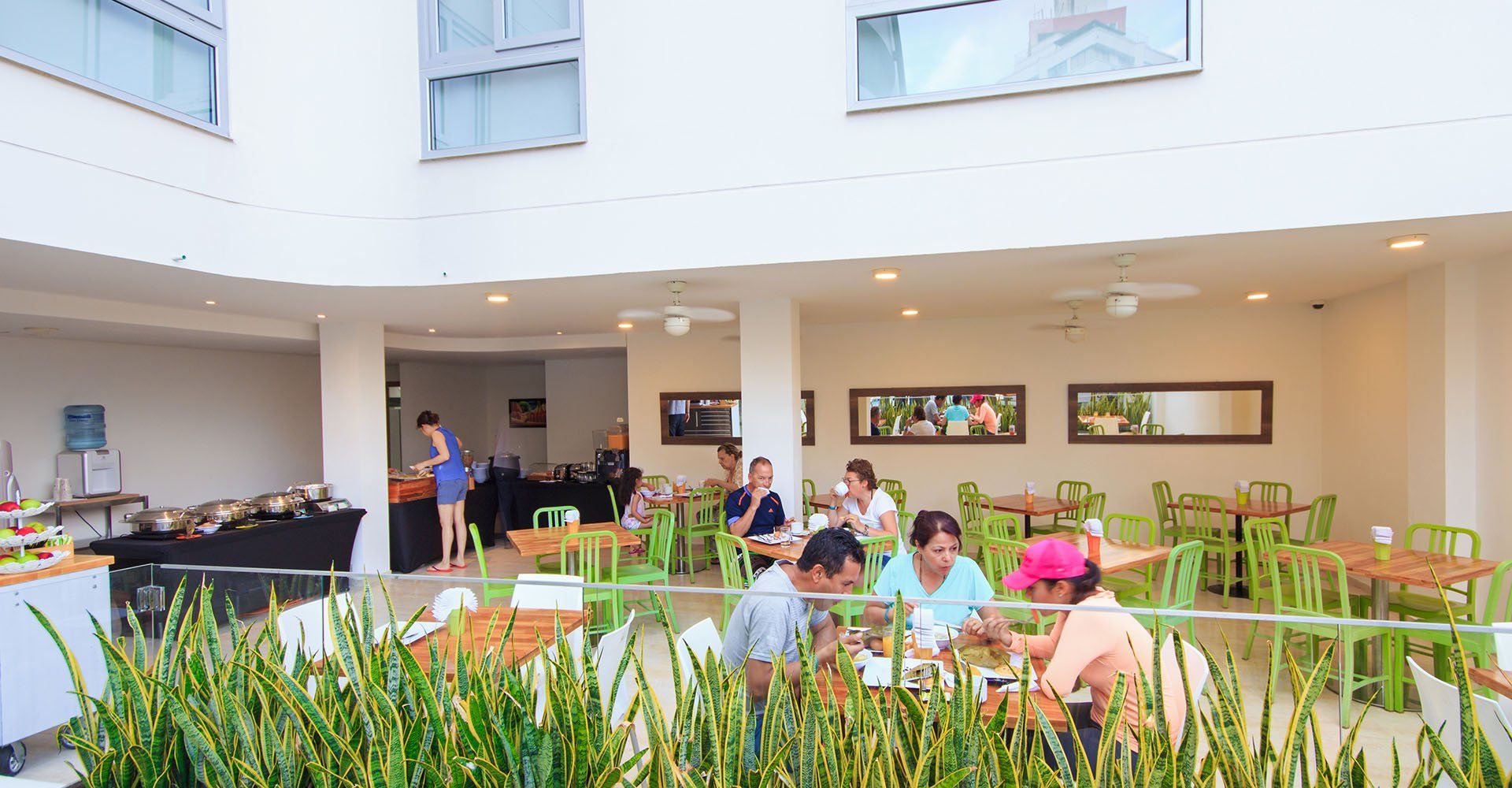 Gente comiendo en mesas al aire libre bajo el patio cubierto de un edificio blanco, con sillas y mesas verdes y una mesa de buffet.