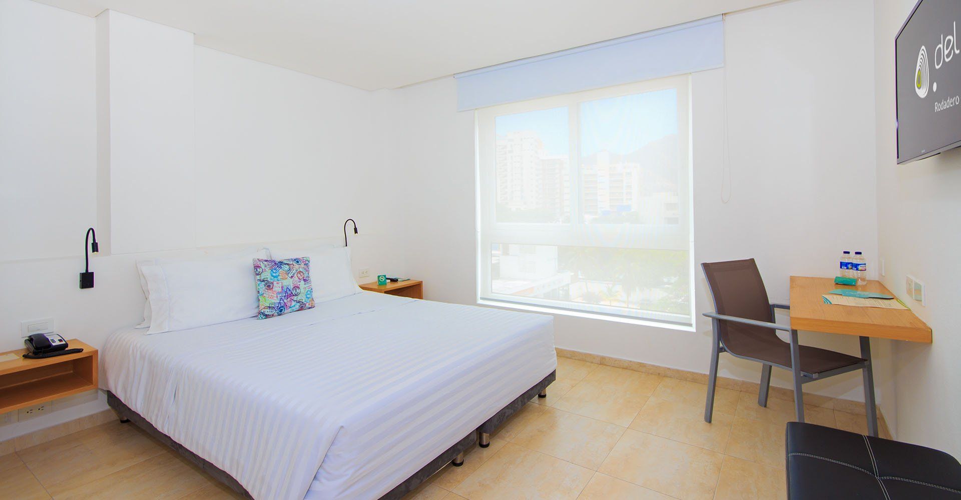 Una habitación de hotel luminosa con cama doble, escritorio y ventana con persianas. Las paredes son blancas.