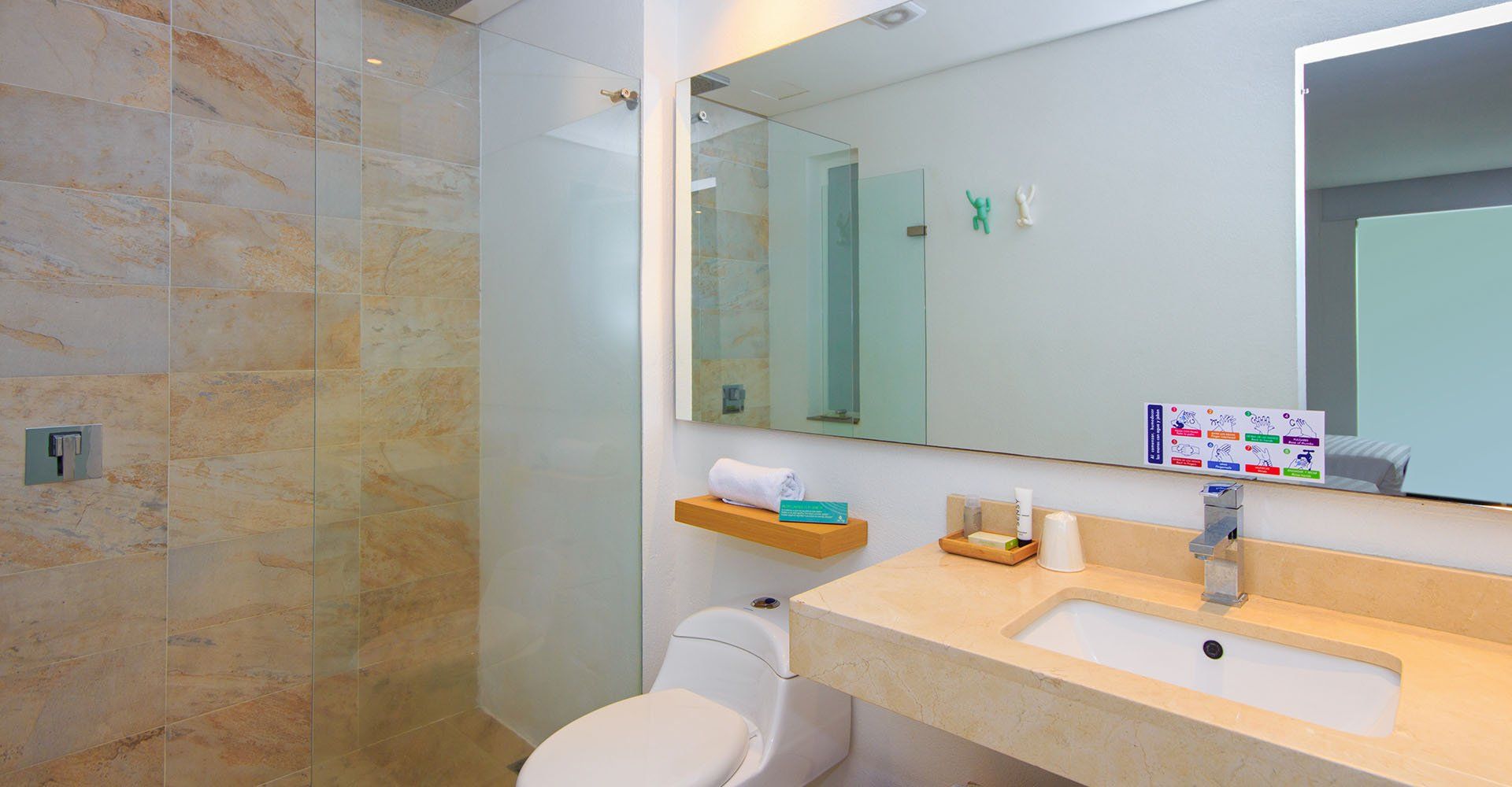 Baño con inodoro blanco, tocador claro con lavabo y espejo grande. Ducha con azulejos beige y estante de madera con artículos de aseo.