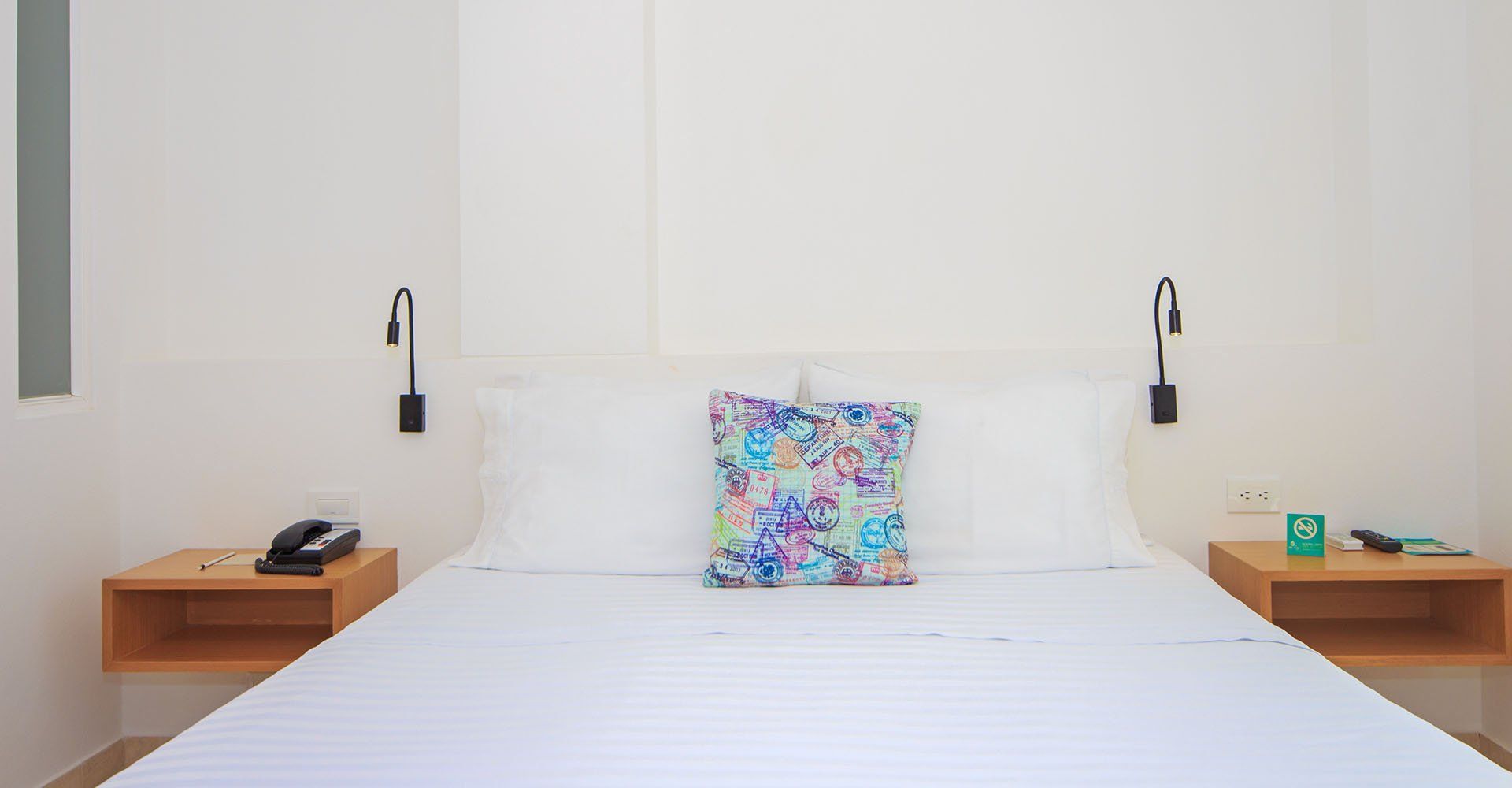 Dormitorio con una cama blanca, dos mesitas de noche de madera y una almohada estampada. Lámparas de noche negras en la pared.