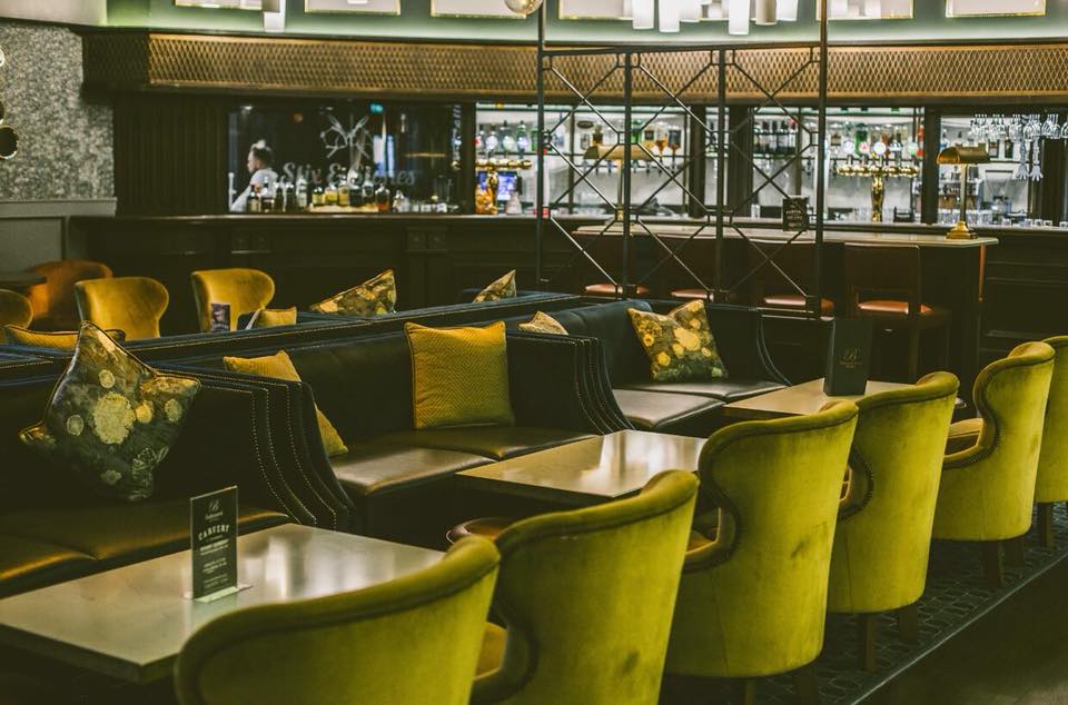 Modern lounge with green velvet chairs, dark tables, and a bar in the background