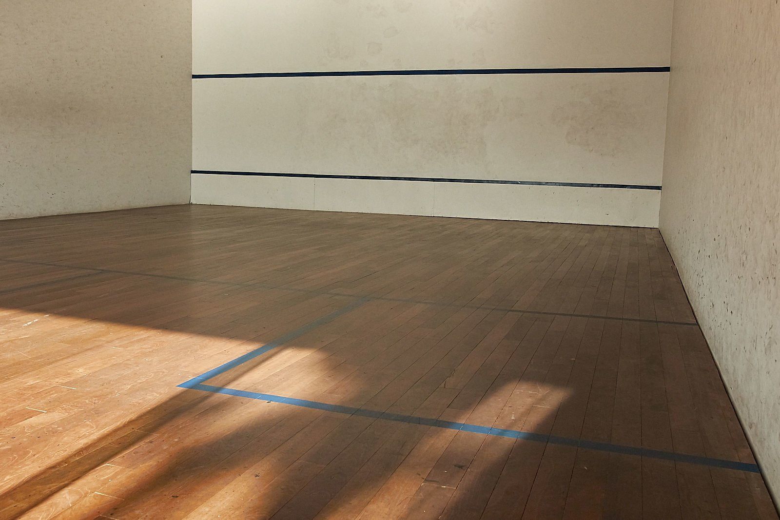 Quadra de squash vazia com piso de madeira marrom e paredes brancas com linhas pretas. A luz do sol projeta sombras no chão.