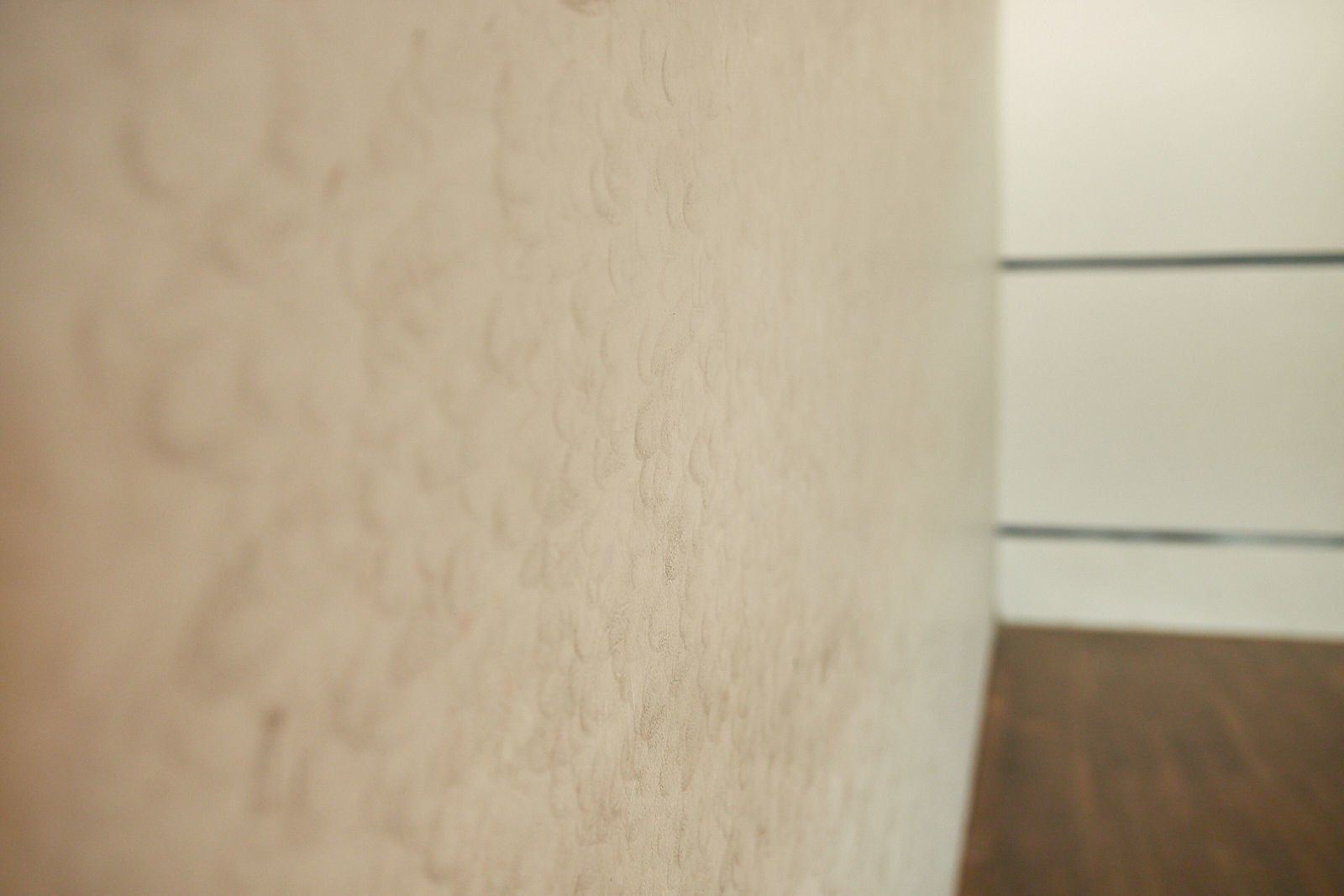 Close-up de uma parede texturizada de cor clara, e um piso escuro são visíveis no fundo desfocado.