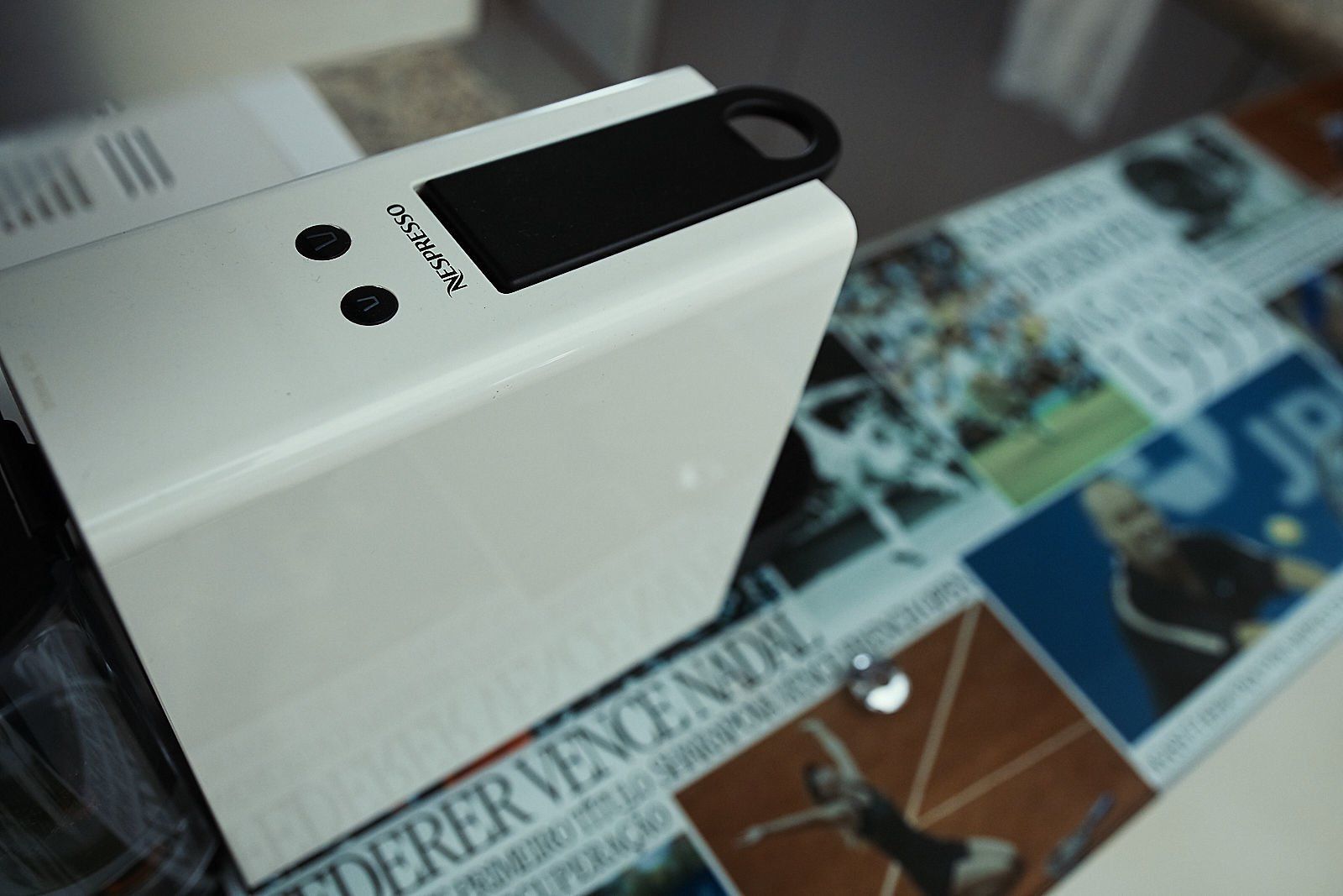Dispositivo eletrônico branco com botões e um laço preto sobre uma mesa de vidro com recortes de notícias.
