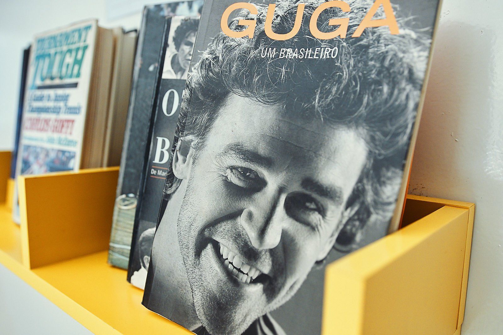 Livros em uma prateleira amarela; uma capa de livro de destaque apresenta foto de um homem sorridente com 