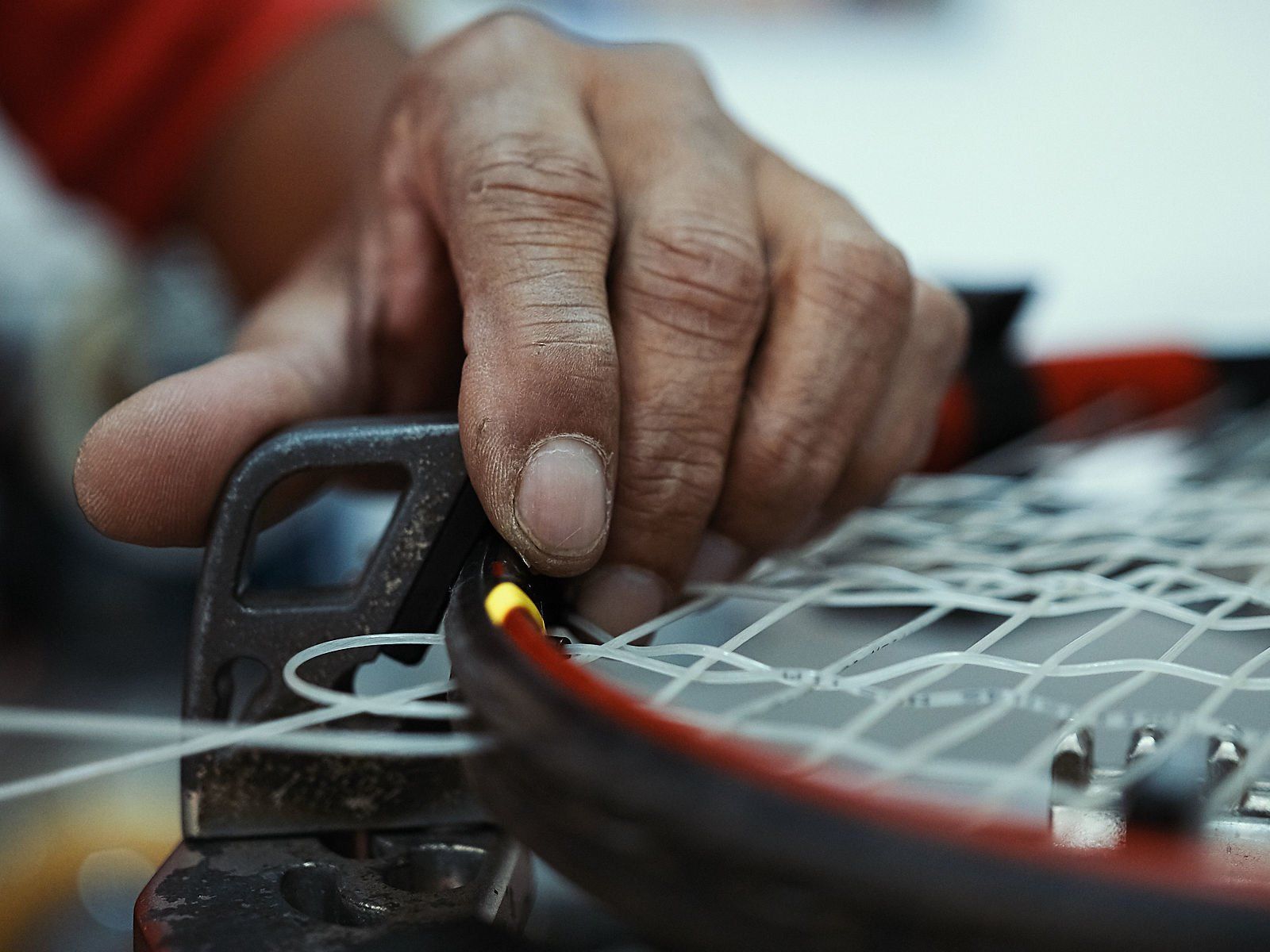 Close da mão de uma pessoa encordoando uma raquete de tênis, usando uma máquina. A estrutura da raquete é preta e vermelha.