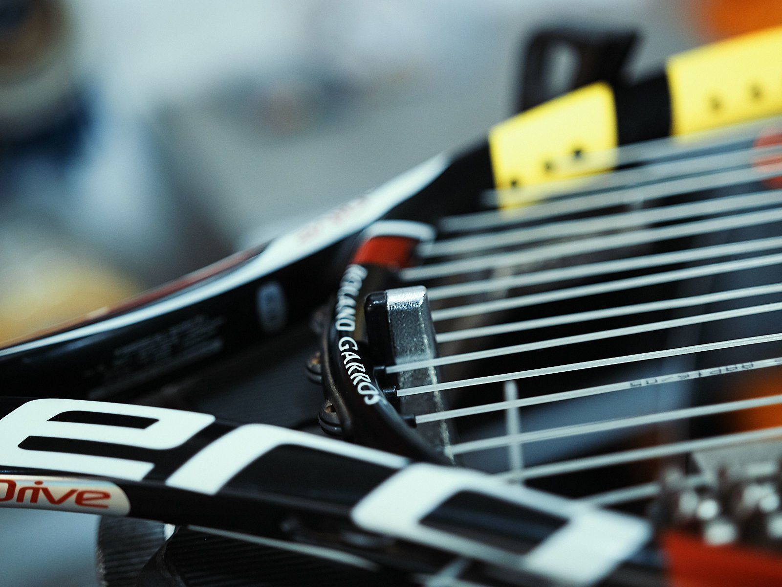 Close-up de uma raquete de tênis com cordas, estrutura preta e branca e marca Roland Garros.