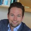 Een man in een blauwe blazer en overhemd lacht, met boekenplanken op de achtergrond.