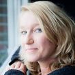 Blonde vrouw met blauwe ogen, leunend tegen een raam, glimlachend.