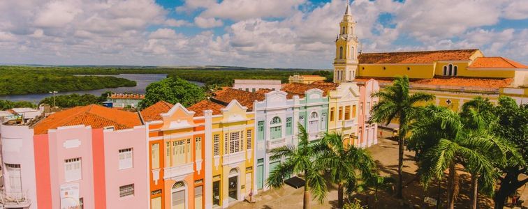 Uma fileira de edifícios coloridos em estilo colonial sob um céu azul, ao lado da torre de uma igreja e de um rio com vegetação exuberante.