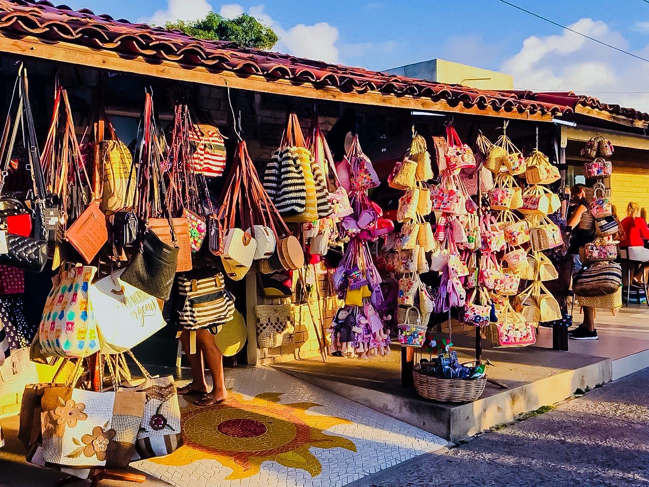 Uma barraca de mercado com um desenho de sol em azulejos exibe uma variedade de bolsas coloridas penduradas e acessórios de palha trançada.