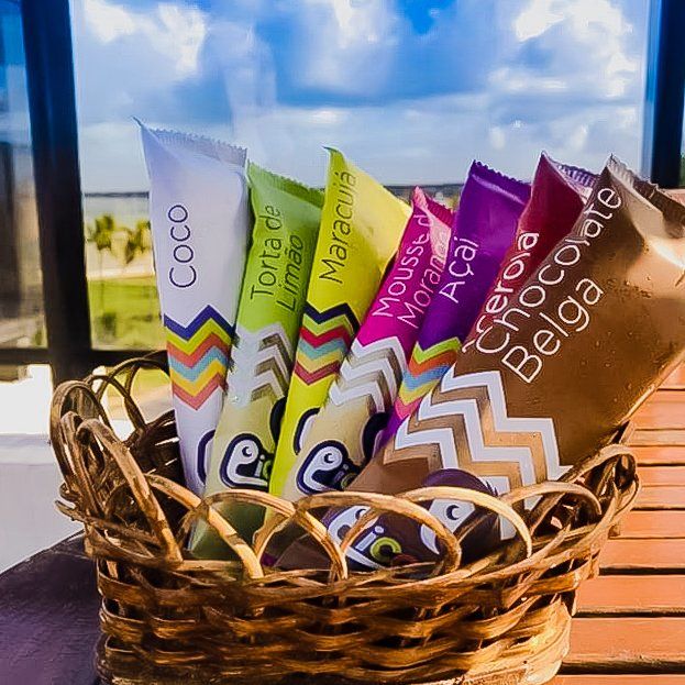 Uma cesta de vime cheia de picolés coloridos de vários sabores, contra um fundo externo desfocado e luminoso.