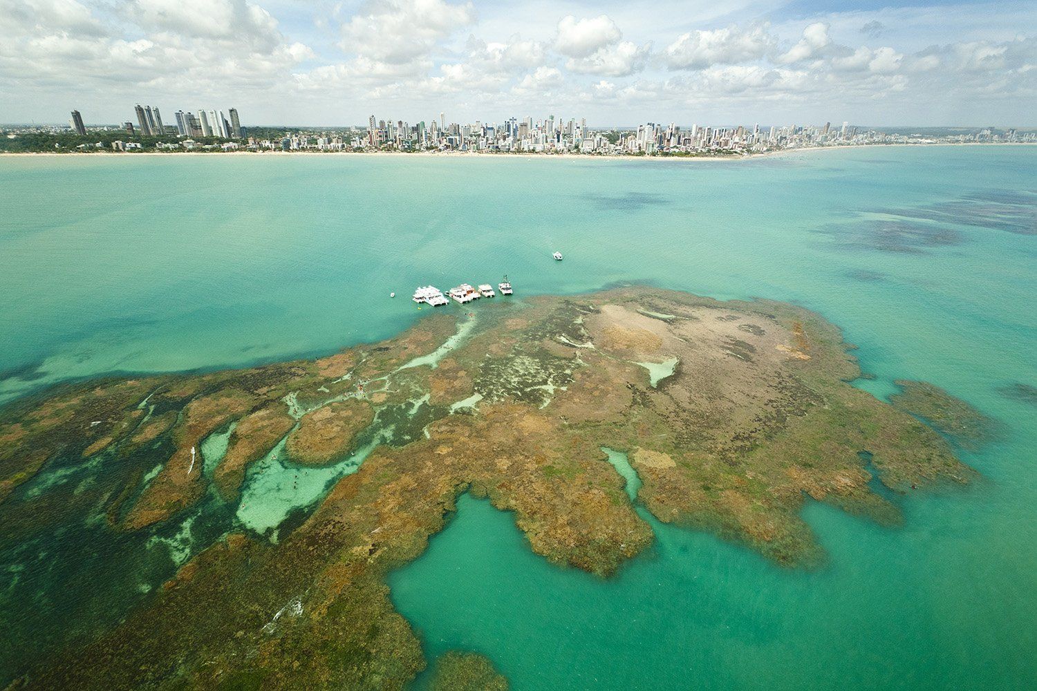 Vista aérea de um recife de coral em águas turquesa, com a costa de uma cidade ao longe e pequenos barcos ancorados perto do recife.