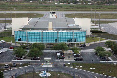 Vista aérea de um terminal aeroportuário moderno com fachada de vidro azul, estacionamentos e torre de controle de tráfego aéreo.