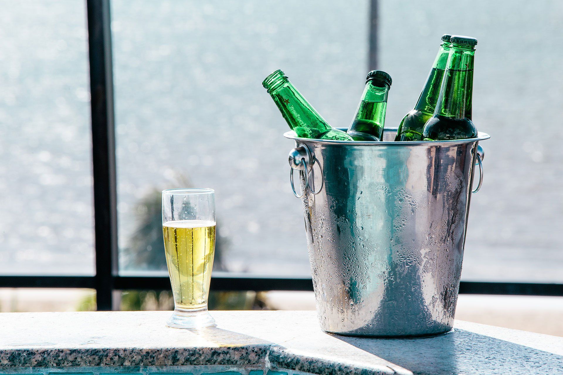 Um balde de gelo contendo quatro garrafas de cerveja de vidro verde, com um copo de cerveja cheio ao lado, em uma prateleira.