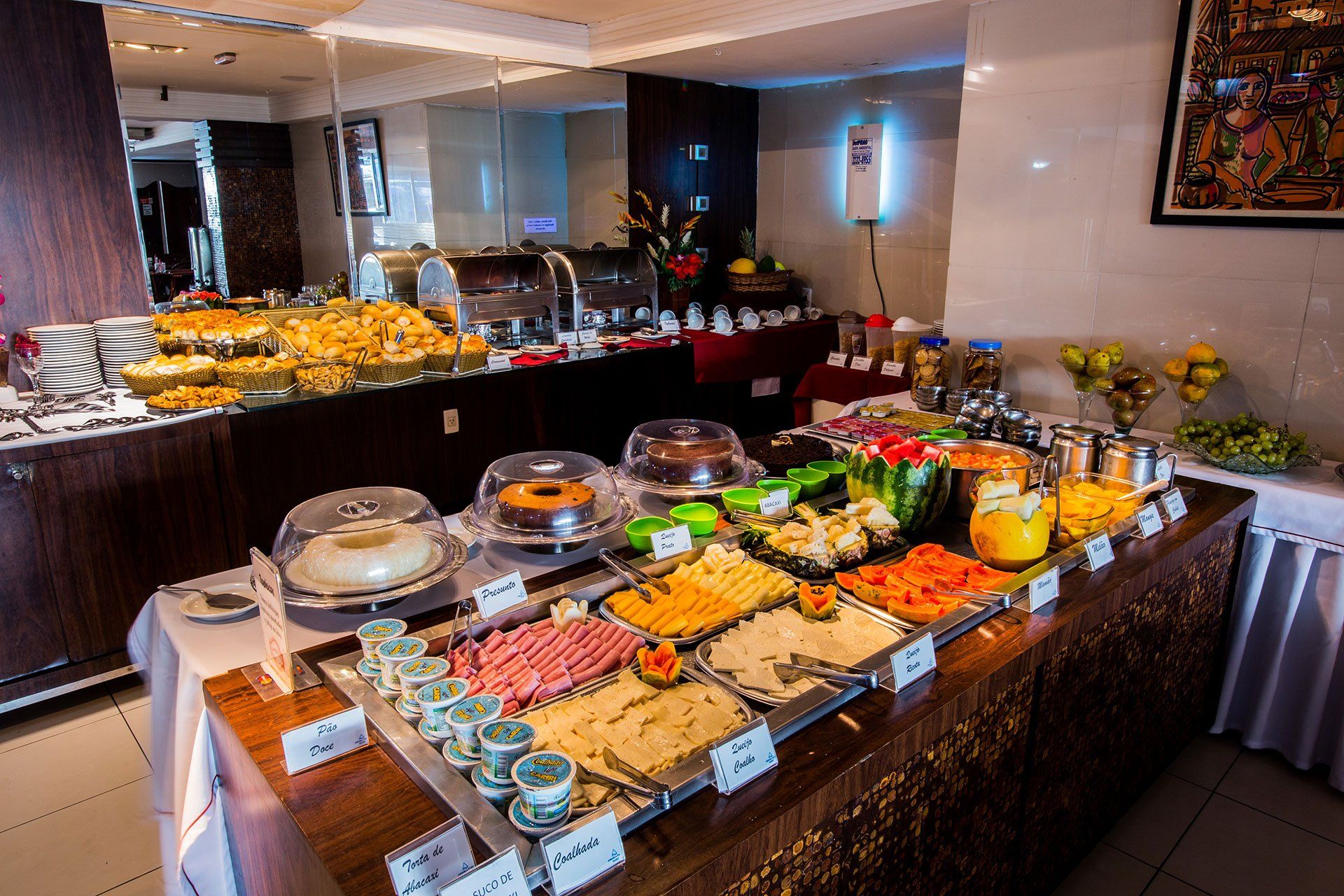 Um buffet de café da manhã de hotel com diversos doces, frutas, frios, queijos e sucos dispostos em uma mesa de madeira.