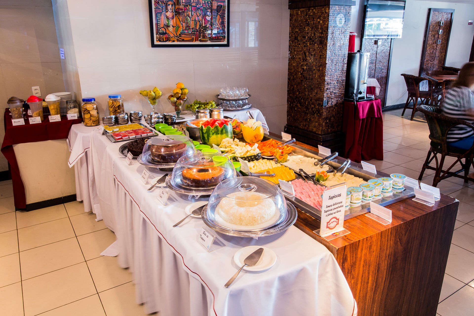 Uma longa mesa de bufê em uma sala de jantar, exibindo uma variedade de bolos, frutas fatiadas, doces e bebidas.