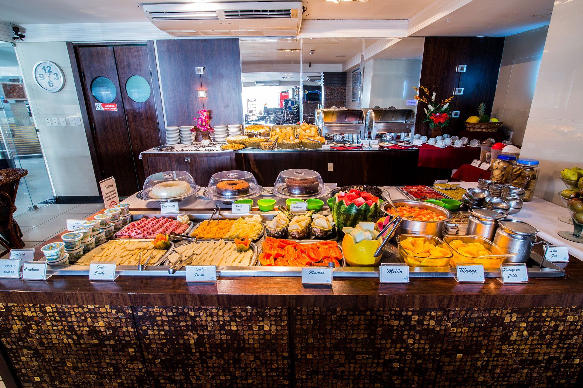 Um buffet de café da manhã de hotel com bandejas de frutas frescas fatiadas, doces e diversos pratos em um balcão de madeira.