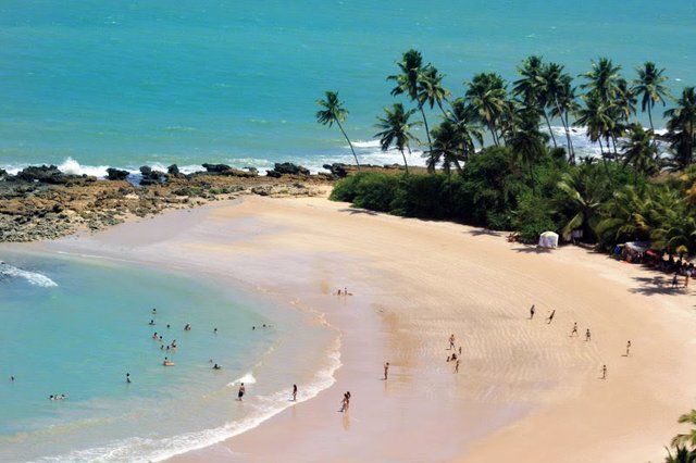 Vista panorâmica de uma praia de areia com palmeiras, formações rochosas ao longo da costa e pessoas nadando e caminhando.