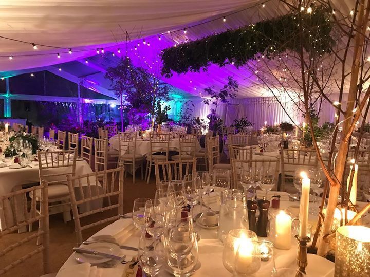 Elegant tented reception with candlelit tables, white chairs, and purple uplighting at dusk