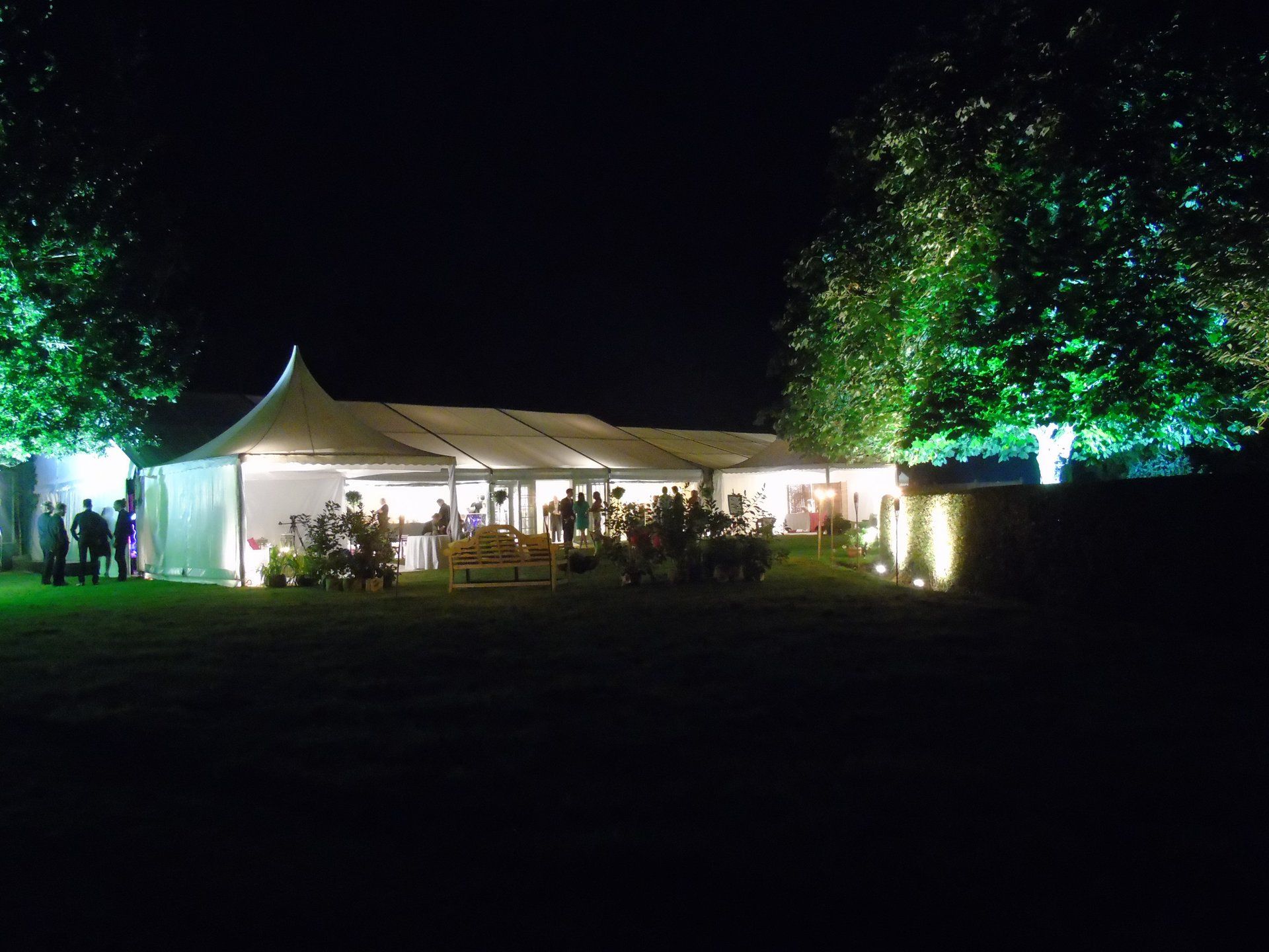 Nighttime outdoor event under a lit white tent, with people gathered and trees illuminated around it.
