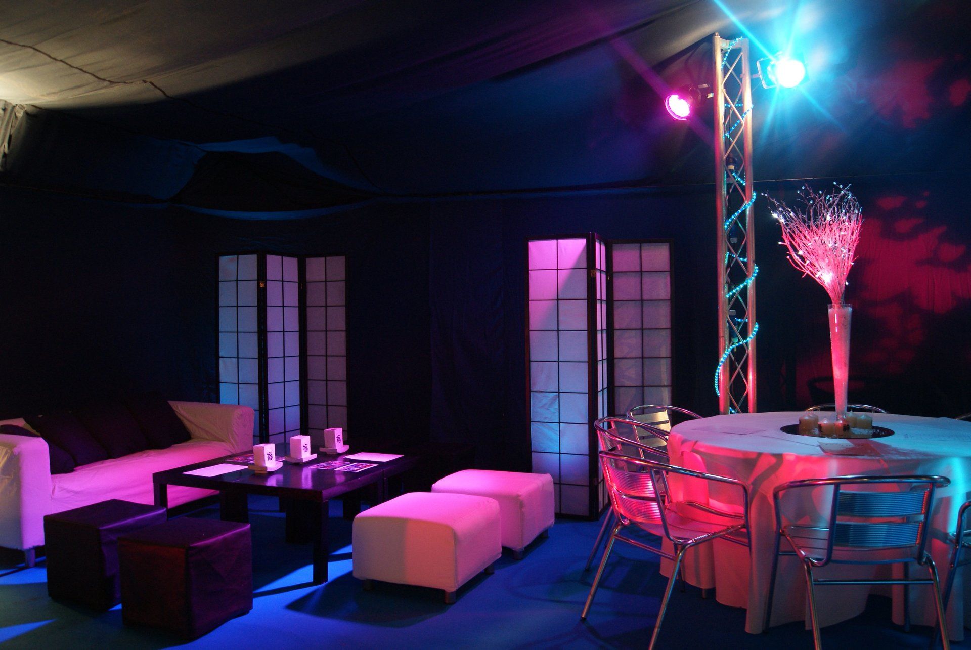 Dim lounge with white sofas, round tables, pink and blue lighting, and decorated cocktail tables under a canopy.