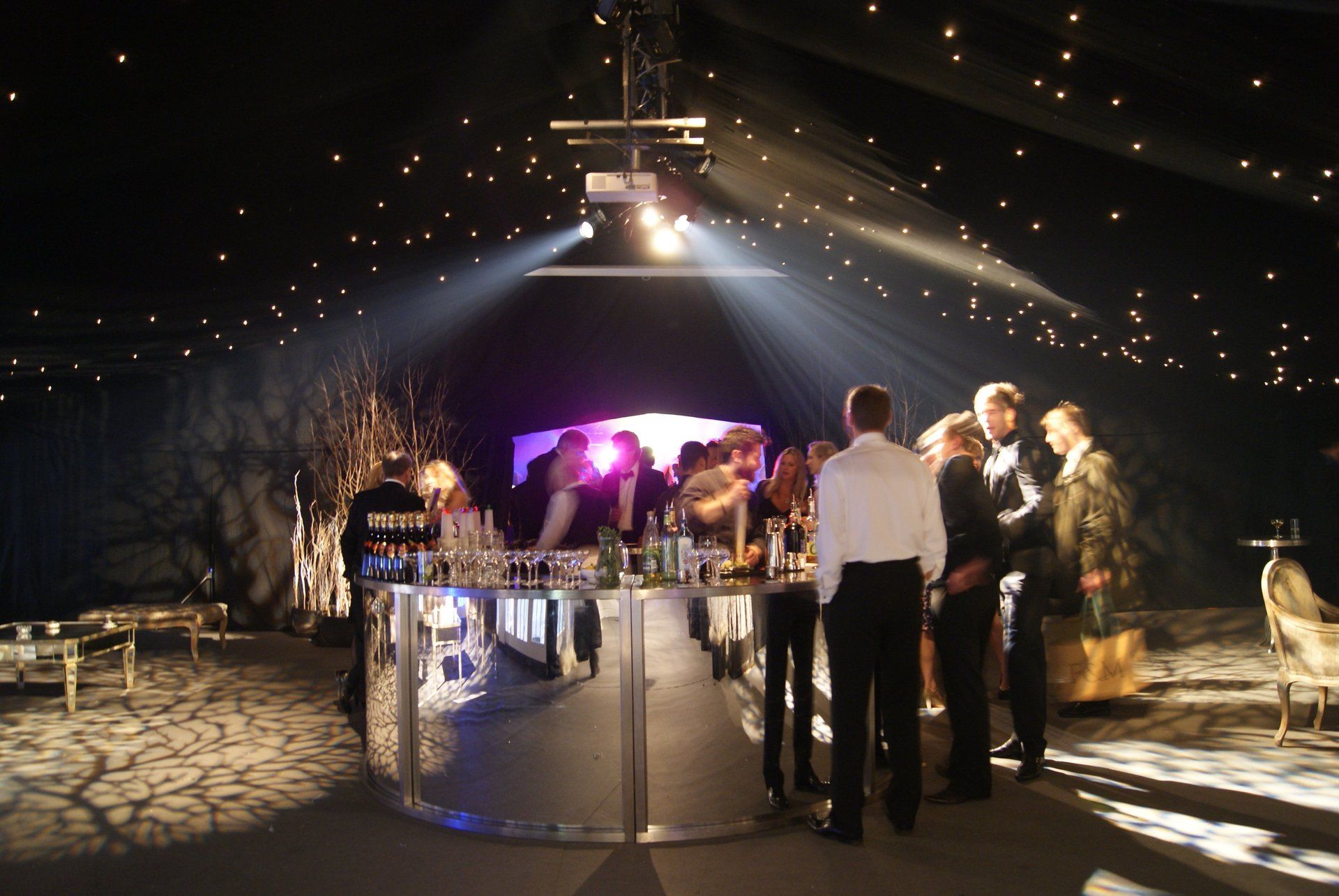 People mingling at a dimly lit event bar under string lights and a spotlighted stage