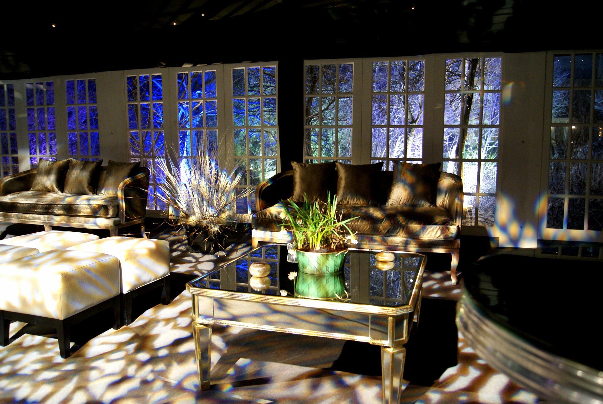 Sunlit patio lounge with wicker sofas, coffee table, and potted plant before blue-lit windows