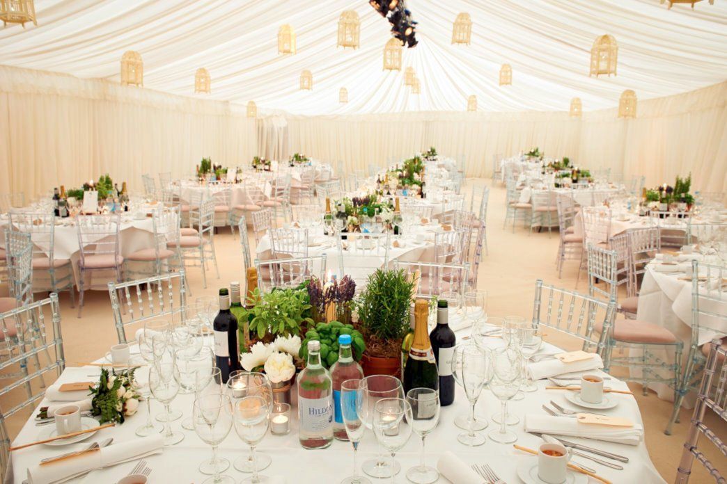 Elegant wedding reception tables under a cream draped tent with gold lights and floral centerpieces