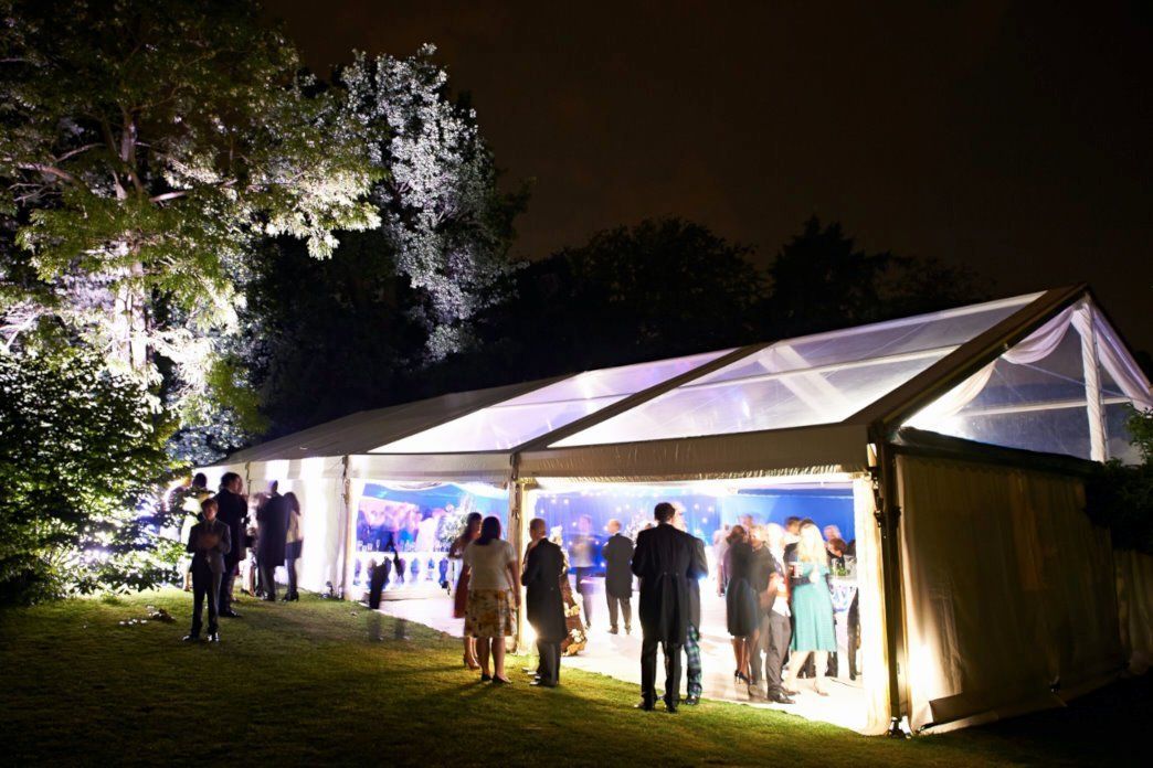 Lit white event tent at night with people gathered on a grassy lawn under trees