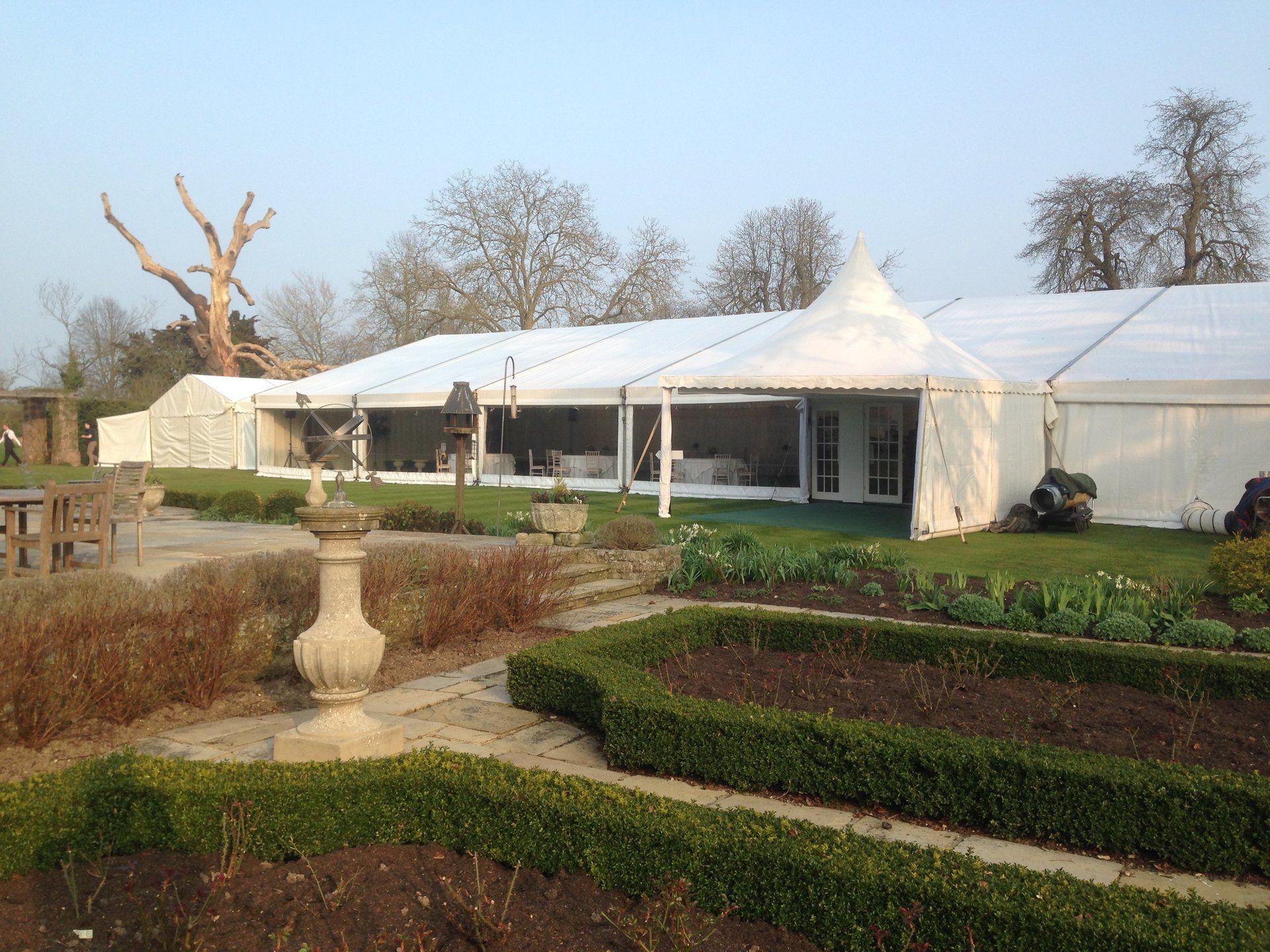 White event tent beside a formal garden with trimmed hedges and a large urn planter under a pale sky