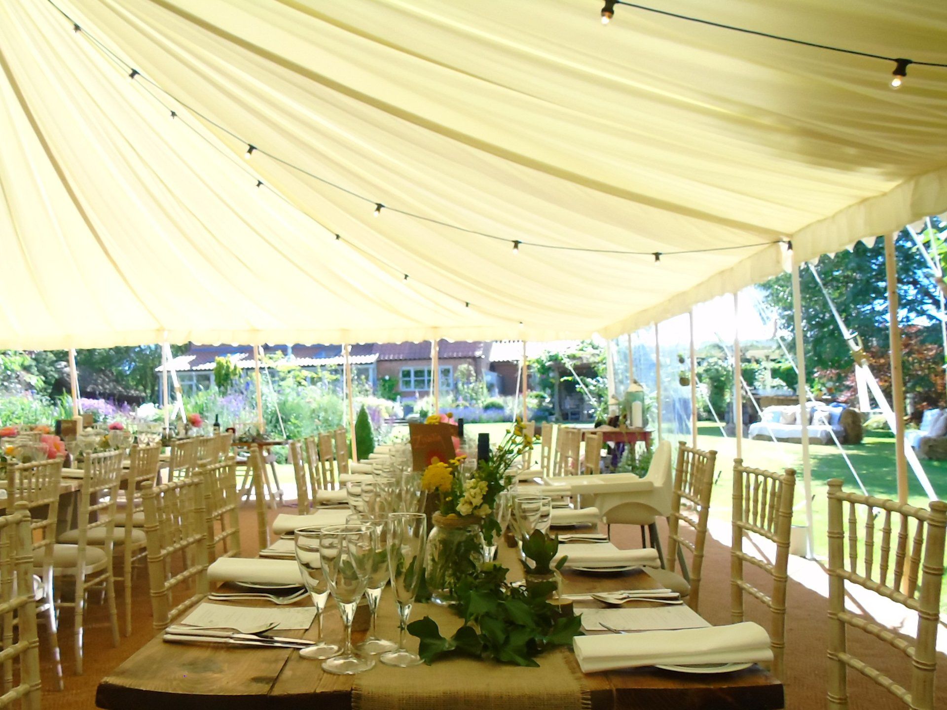 Outdoor banquet tent with cream drapes, wooden tables, white chairs, and garden views