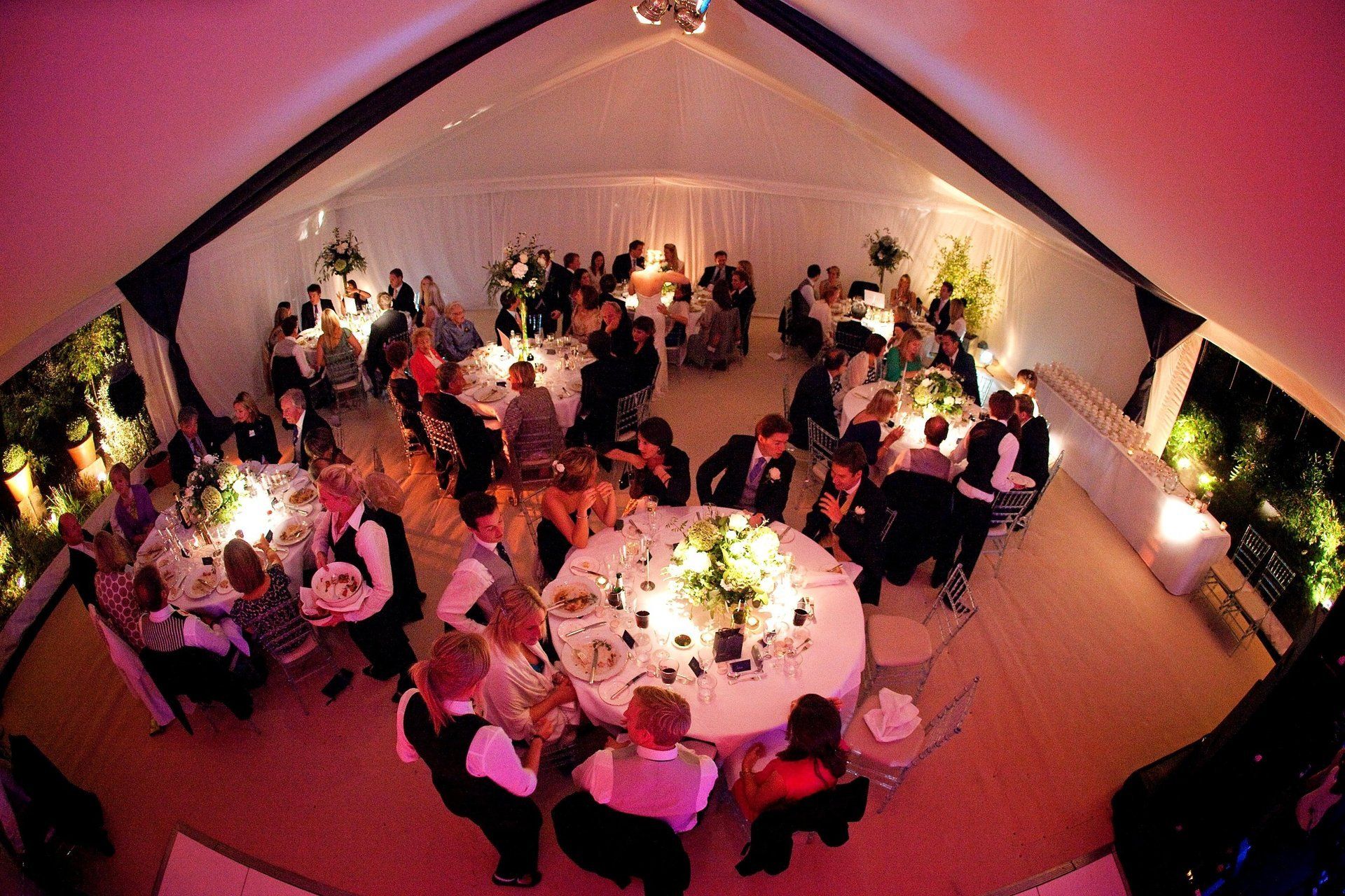 Guests mingling at a formal dinner inside a pink-lit tent with round tables and candles.