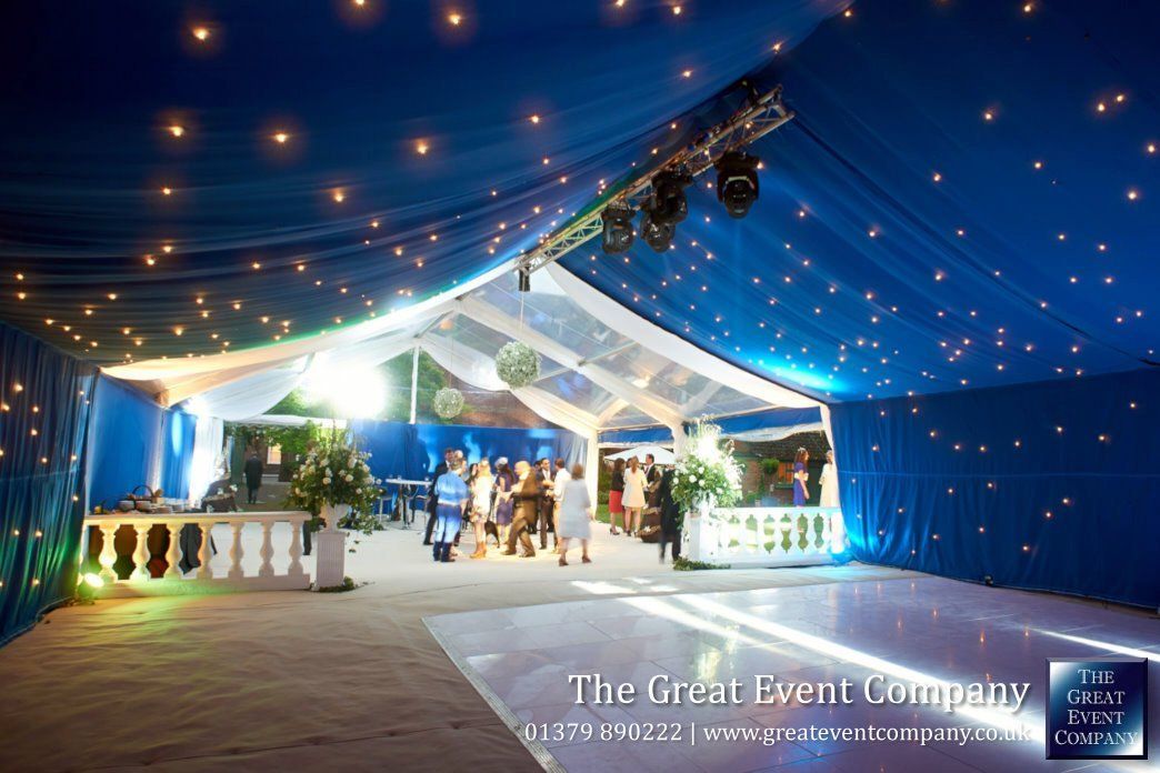 Outdoor wedding reception under blue draped tents with string lights and guests gathered near a decorated stage